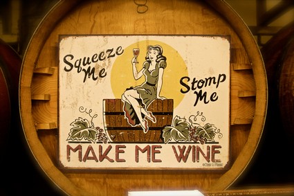 Vintage sign with pin-up style woman holding a wine glass, promoting wine making with the phrase "Squeeze Me, Stomp Me, Make.