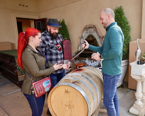People enjoying wine tasting outdoors at Lodi Winegrape Commission with wine barrel and greenery in background.