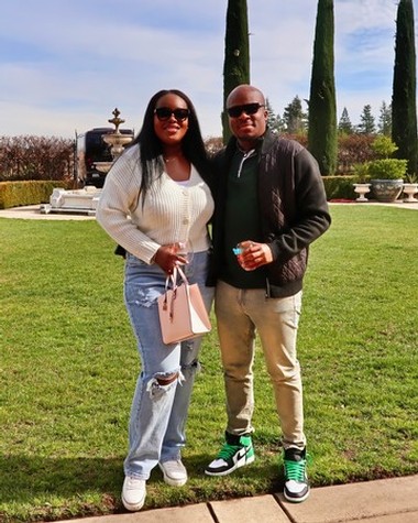 Two people standing outdoors with tall cypress trees and a fountain in the background.