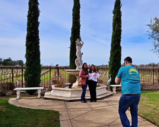 Tourists taking photos at a vineyard with a classical fountain and tall cypress trees in Lodi, California.