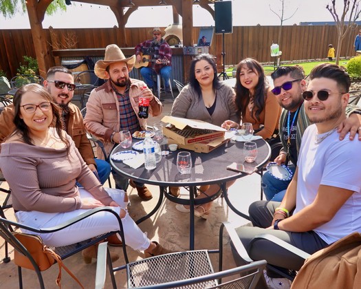 Friends sitting around a table outdoors, smiling, with drinks and food, in a backyard setting.