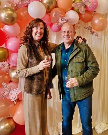 Two smiling people at a celebration with pink, white, and gold balloons, holding wine glasses.