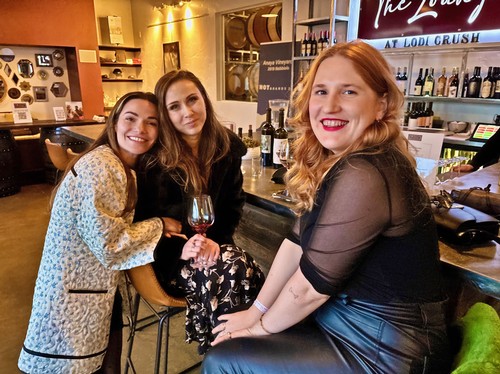 Three women enjoying wine at a bar, smiling and socializing, representing Lodi Winegrape Commission activities.