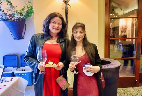 Two women at a wine event holding glasses of wine and a plate of cheese, smiling at the camera.