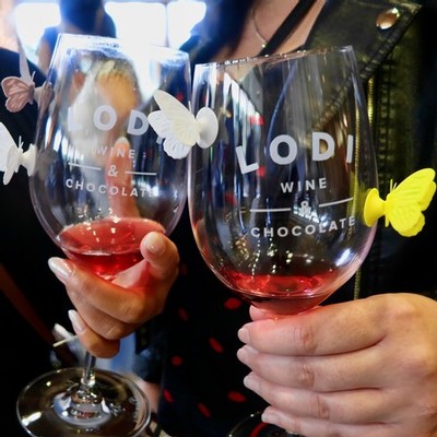 Two wine glasses with Lodi Wine logo, one with red wine, held by people at a tasting event.