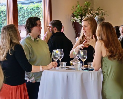 Group of people engaging in conversation at a wine tasting event, with wine glasses on a table.