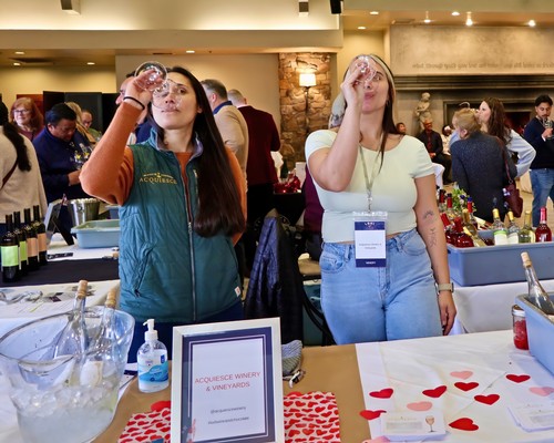 Two women tasting wine at the Lodi Winegrape Commission event, surrounded by wine bottles and attendees.