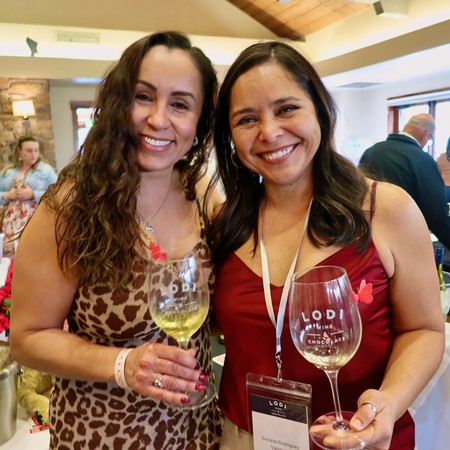 Two women smiling at a wine event, holding glasses of white wine, with a lively background.