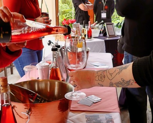 People pouring wine at a tasting event with wine bottles and glasses on a table.