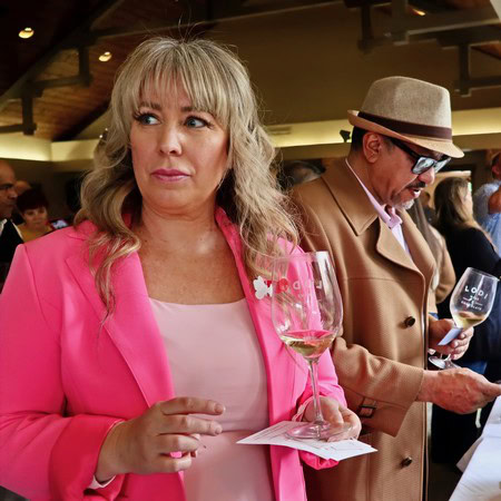 Woman tasting wine at Lodi Winegrape Commission event, holding a glass of white wine in a lively indoor setting.