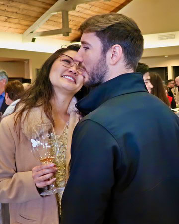 A woman and man sharing a kiss at a Lodi Winegrape Commission event, holding a glass of wine, in a lively indoor setting.
