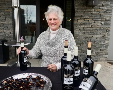 Elderly woman with white hair smiling at a wine tasting event with bottles of wine on the table.