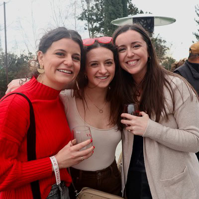 Three women smiling at a wine tasting event, holding glasses, outdoors with trees in the background.