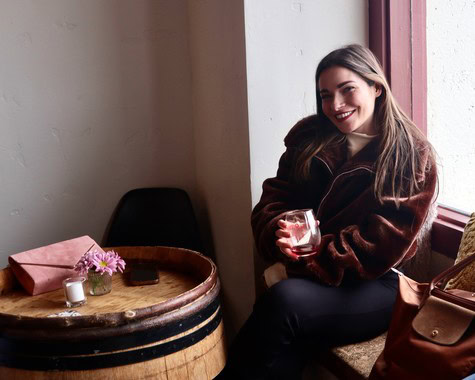 Smiling woman enjoying wine at Lodi Winegrape Commission event, sitting by window with glass of wine.