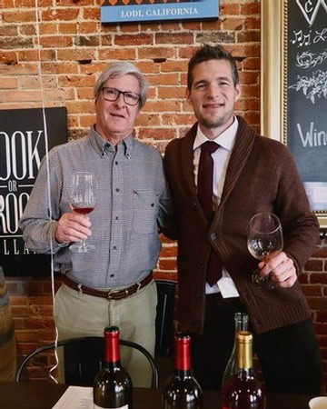 Two men holding wine glasses at Lodi Winegrape Commission event in front of brick wall.