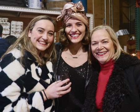 Three women smiling at a wine event, holding a glass of red wine, in a warehouse setting.