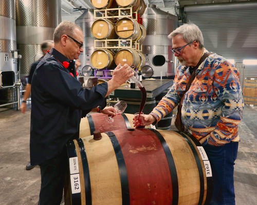 Two men tasting wine from a barrel at a winery, with wine barrels in the background.