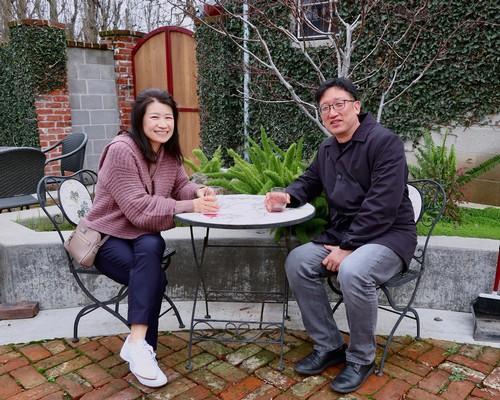Two people sitting at a table outdoors in a garden, smiling, with lush greenery and brick paving.