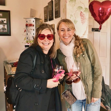 Two women smiling at a wine event, holding glasses of red wine, with a heart-shaped balloon in the background.