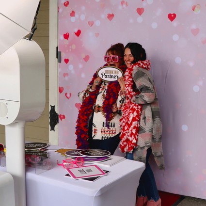 Two women at Lodi Winegrape Commission event with heart-themed backdrop and photo props.