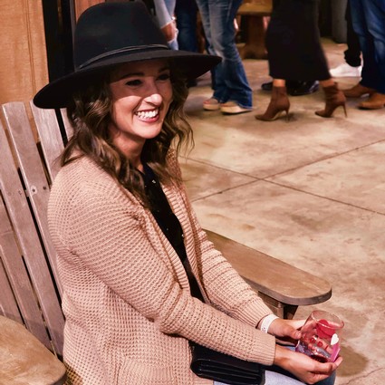Smiling woman in a black hat and beige sweater at a wine event, holding a glass of wine.