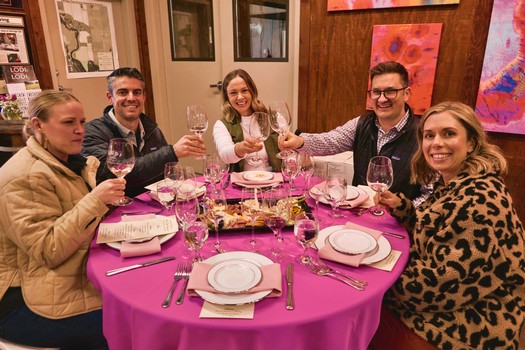 Group of five people enjoying wine and dinner at a round table with pink tablecloth, smiling and toasting.