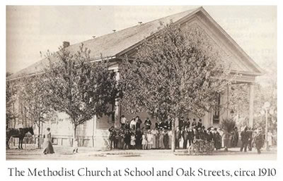 Historic black and white photo of the Methodist Church at School and Oak Streets circa 1910 with people and horse-drawn carra.