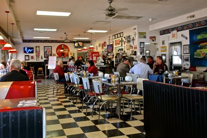 Interior of a lively restaurant with patrons enjoying meals and drinks, decorated with wine-themed artwork and memorabilia.
