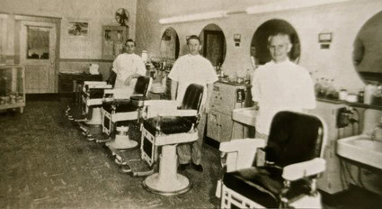 Black and white photo of a vintage barber shop with four barbers and customers.