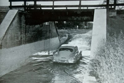 Old vintage car submerged in water under a bridge, black and white photo, historical scene.