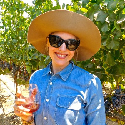 Woman in vineyard with wide-brim hat and sunglasses holding a glass of wine.