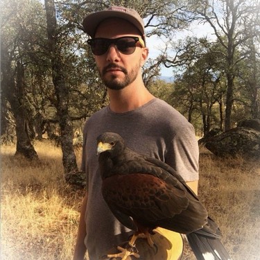 Man wearing sunglasses and cap holding a bird on his arm in a natural outdoor setting.