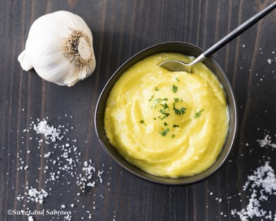 Creamy mashed potatoes garnished with herbs in a black bowl, with garlic and salt on a dark wooden surface.