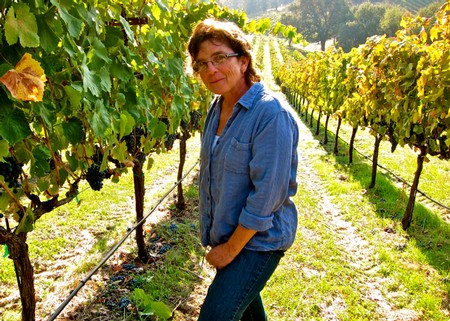 Woman in a blue shirt standing in a lush vineyard with grapevines on both sides.