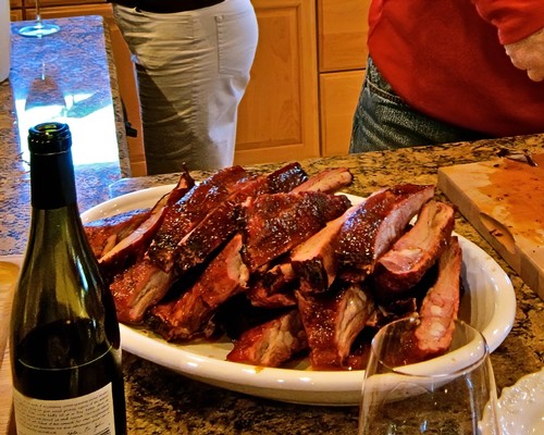 Barbecue ribs with wine on a kitchen counter, people in background, warm indoor lighting.