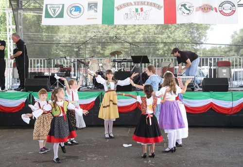 Children in traditional costumes performing at a cultural event with a stage and musical instruments in the background.