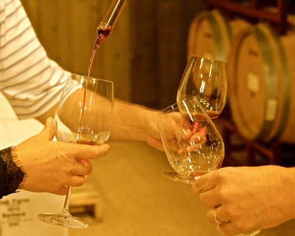 People tasting wine at a winery, pouring wine into glasses, wine barrels in the background.