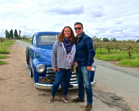 Two people standing in front of a vintage blue truck in a vineyard, smiling at the camera.