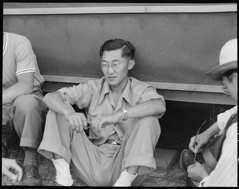 Asian man sitting on ground, wearing glasses and casual clothing, engaged in conversation or listening.