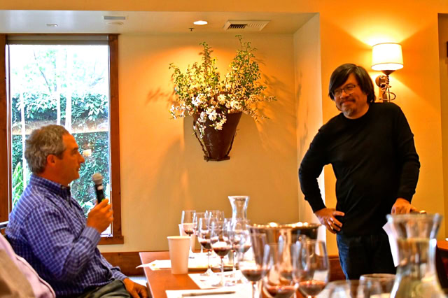 Two men in a wine tasting event, one speaking into a microphone, the other listening, with wine glasses on the table.