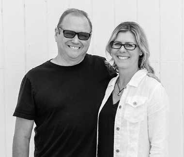 Smiling man and woman wearing glasses, standing together outdoors in black and white.
