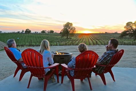 Group of people enjoying a sunset over a vineyard in Lodi, California, with red chairs and a fire pit.