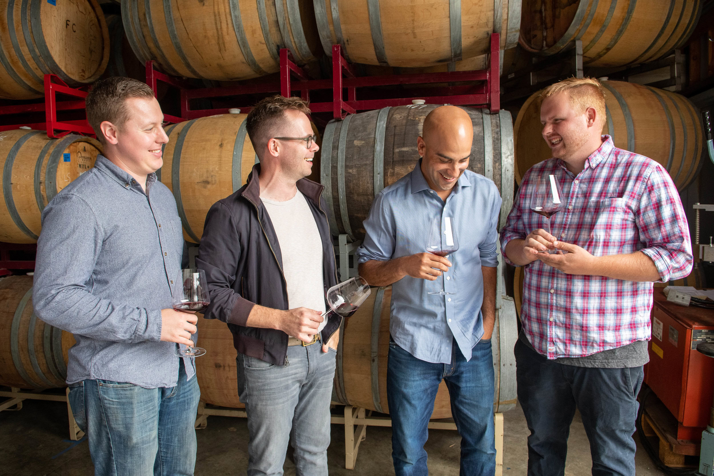 Group of men tasting wine in a winery with wine barrels in the background.