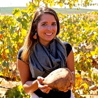 Smiling woman holding a large grape in a vineyard with lush green vines in the background.