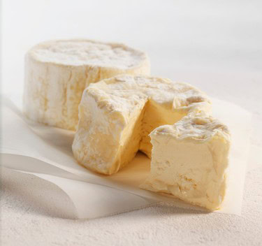 Close-up of a soft, creamy cheese wheel with a slice cut out, on a white surface.