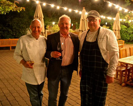 Three men at an outdoor wine event, smiling and holding wine glasses, under string lights at night.
