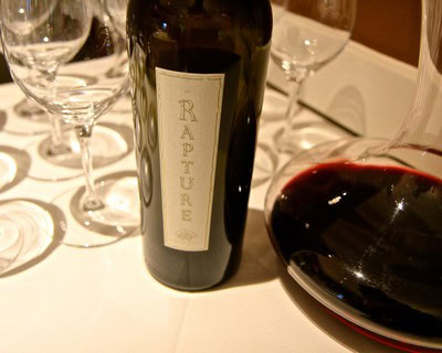 Close-up of a wine bottle labeled 'Rapture' on a table with wine glasses.