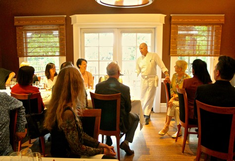 Chef presenting to a group of people in a cozy restaurant setting with large windows and wooden blinds.