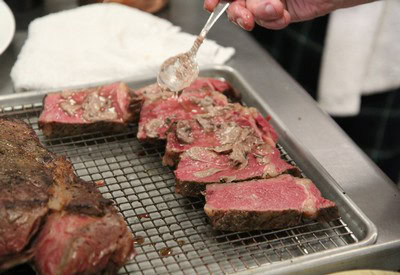 Chef drizzling sauce over cooked beef slices on a wire rack in a professional kitchen.