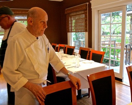 Chef in a white uniform preparing a table in a cozy dining room with large windows and wooden furniture.